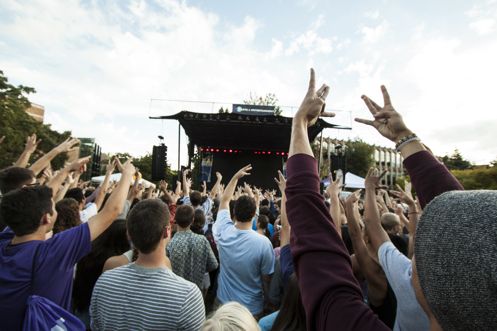 A crowd of young adults stand in front of a stage with their hands raised up in the air.