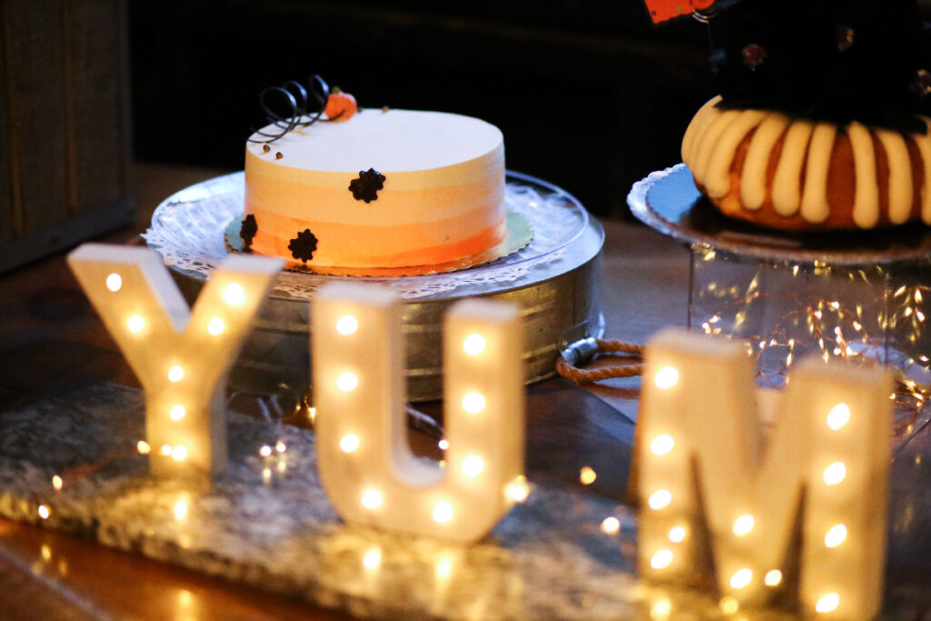 A frosted cake on a table sits next to a brightly-lit "YUM" sign.