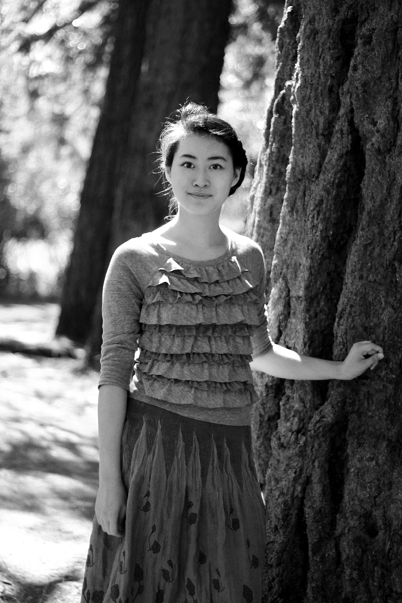 A young woman stands nearby a tree in the forest, staring into the camera with a small smile, with her left hand resting on the trunk.