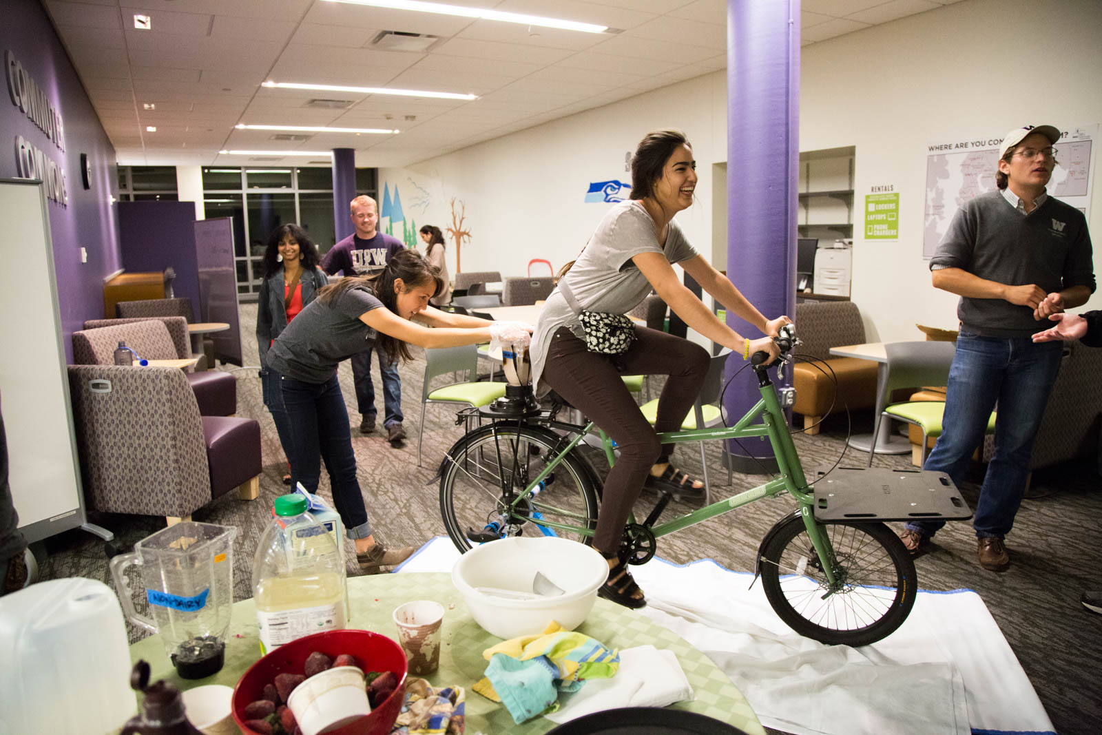 Inside a University of Washington building, a student rides on a bicycle with a blender attached to the back.