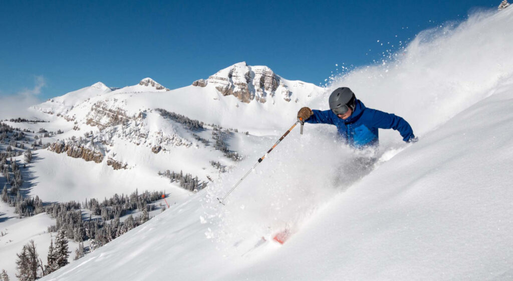 A skier wearing a coat and helmet goes down a mountain, with fresh snow powder all around them.
