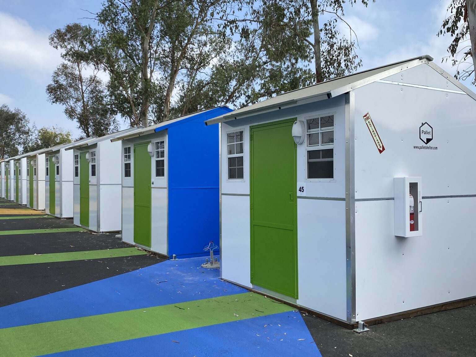 A row of tiny houses (shelters) built by Pallet Shelter. Each has a colorful front door and two windows.