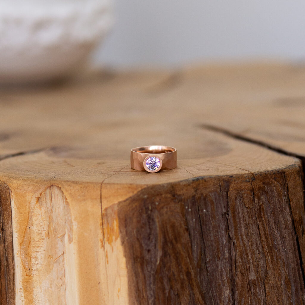 A rose gold banded ring with a center diamond sits on a log table.
